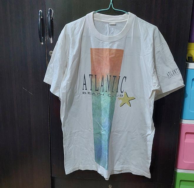 เสื้อยืดเป็นเสื้อวินเทจสีขาวลายกราฟิกด้านหน้าพร้อมตัวอักษร "ATLANTIC BEACH CLUB