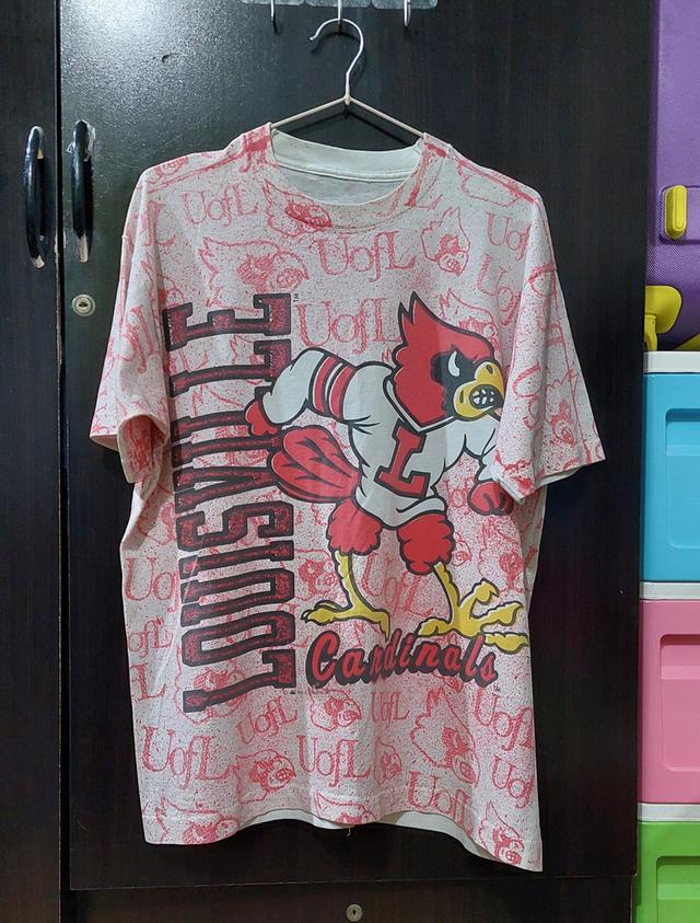 เสื้อยืดวินเทจลาย Louisville Cardinals AOP (All Over Print)จากช่วงยุค90sมีลายพิมพ์แบบเต็มตัวและตัวมาสคอตนกคาร์ดินัลที่เป็นสัญลักษณ์ของทีม โลโก้ "UofL