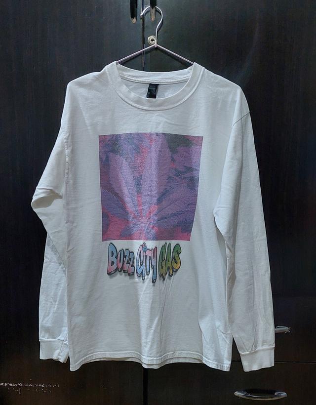 เสื้อยืดแขนยาวสีขาว มีสกรีนลายกราฟิกรูปใบพืชขนาดใหญ่ในกรอบสี่เหลี่ยมสีสันแนวไซเคเดลิก (psychedelic) และมีข้อความ "Buzz City GAS" อยู่ด้านล่าง รูปย่อยที่ 1